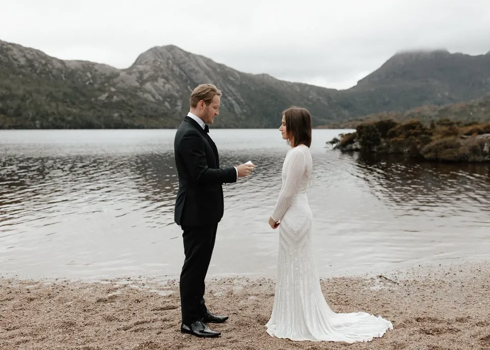 Josh reading from his vow booklet as April listens on the lakeshore, at Dove Lake, Cradle Mountain, Tasmania, for an Elopement Collective elopement. Photo by Bulb Creative.