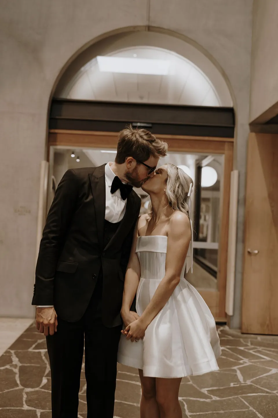 Danielle and Adam kissing beneath a stone arch in the hotel corridor, at The Calile Hotel in Fortitude Valley, Brisbane, for an Elopement Collective elopement. Photo by George Bowden.