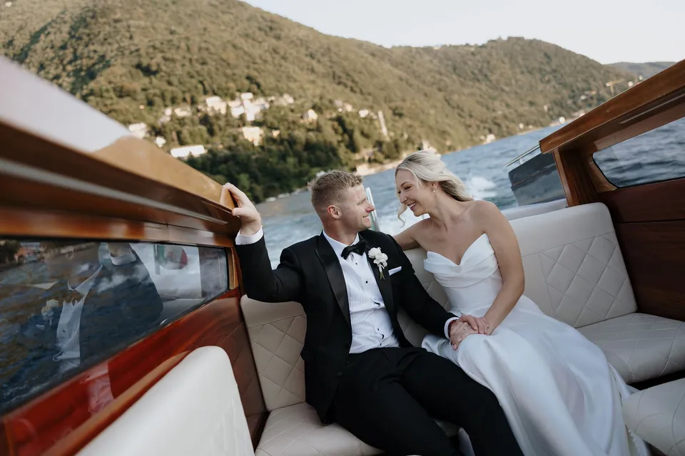 Zoe and Charlton laughing together on a white boat seat, with Charlton’s hand resting on the wooden window frame, on Lake Como in Lombardy, Italy, for an Elopement Collective elopement. Photo by George Bowden.