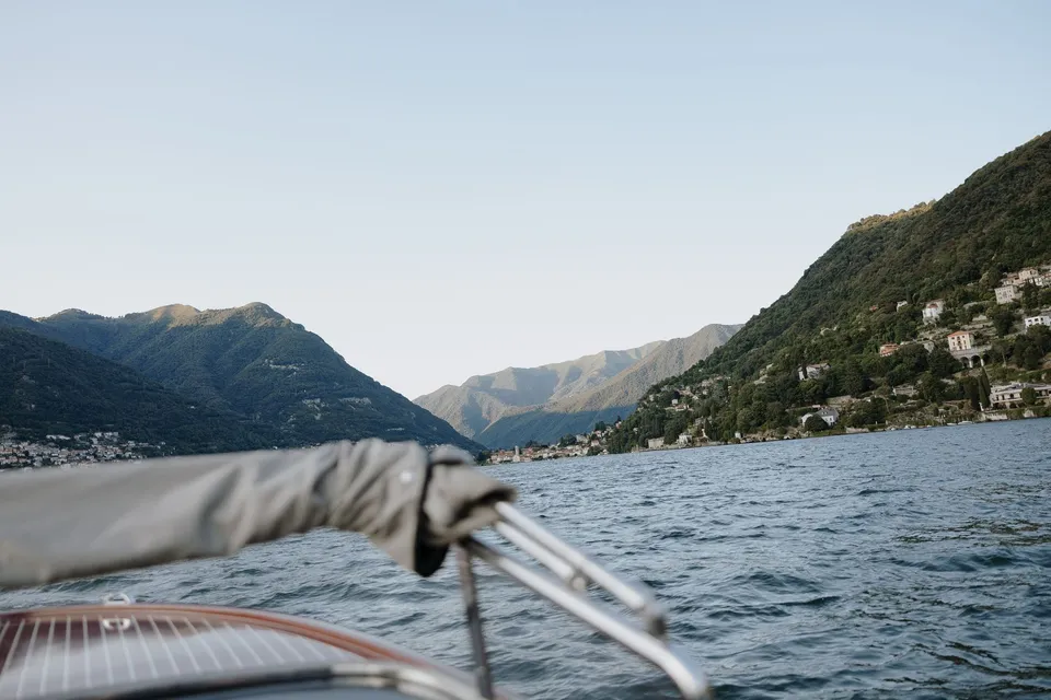 View over the bow of a boat towards blue water, lakeside villages and mountain ridges, on Lake Como in Lombardy, Italy, for an Elopement Collective elopement. Photo by George Bowden.
