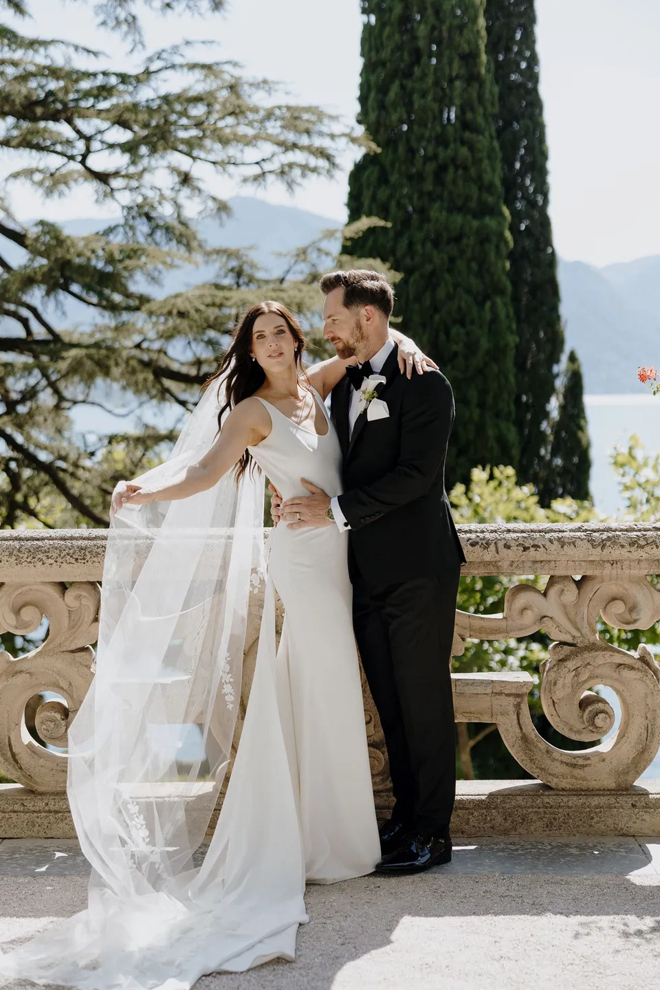 Lachlan embracing Lyndal as she lifts the edge of her embroidered veil beside a carved stone balustrade on Villa del Balbianello’s lakeside terrace, at Lake Como in Lombardy, Italy, for an Elopement Collective elopement. Photograph by George Bowden.