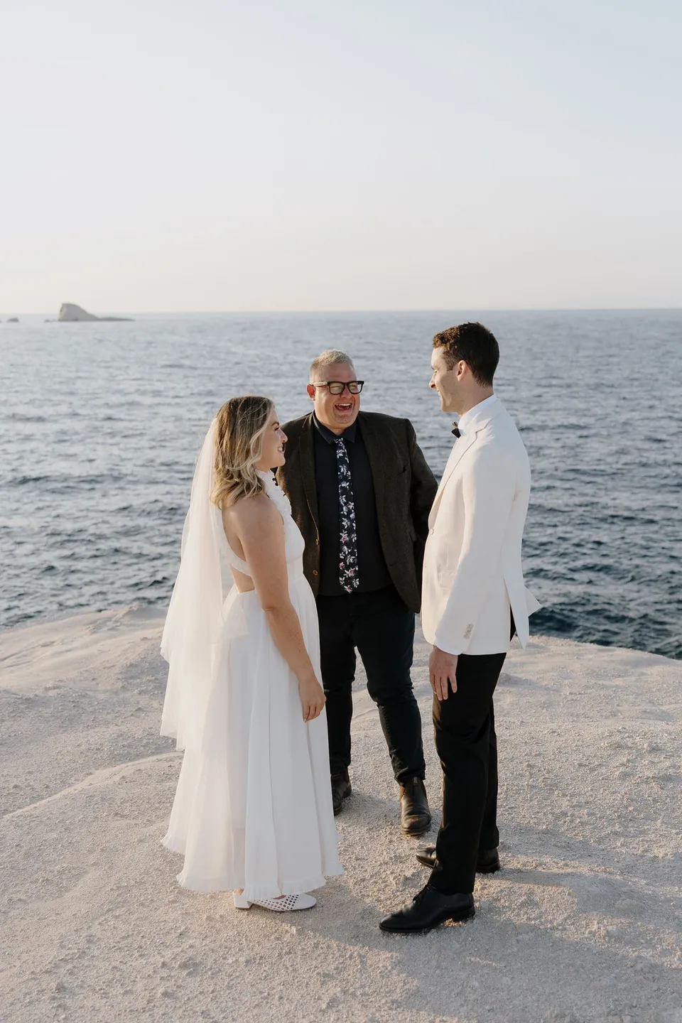 Georgina and Brad facing each other during their ceremony, with celebrant Josh Withers between them and the rocky coastline behind, at Sarakiniko on Milos in the Cyclades, Greece, during an Elopement Collective elopement. Photograph by George Bowden.