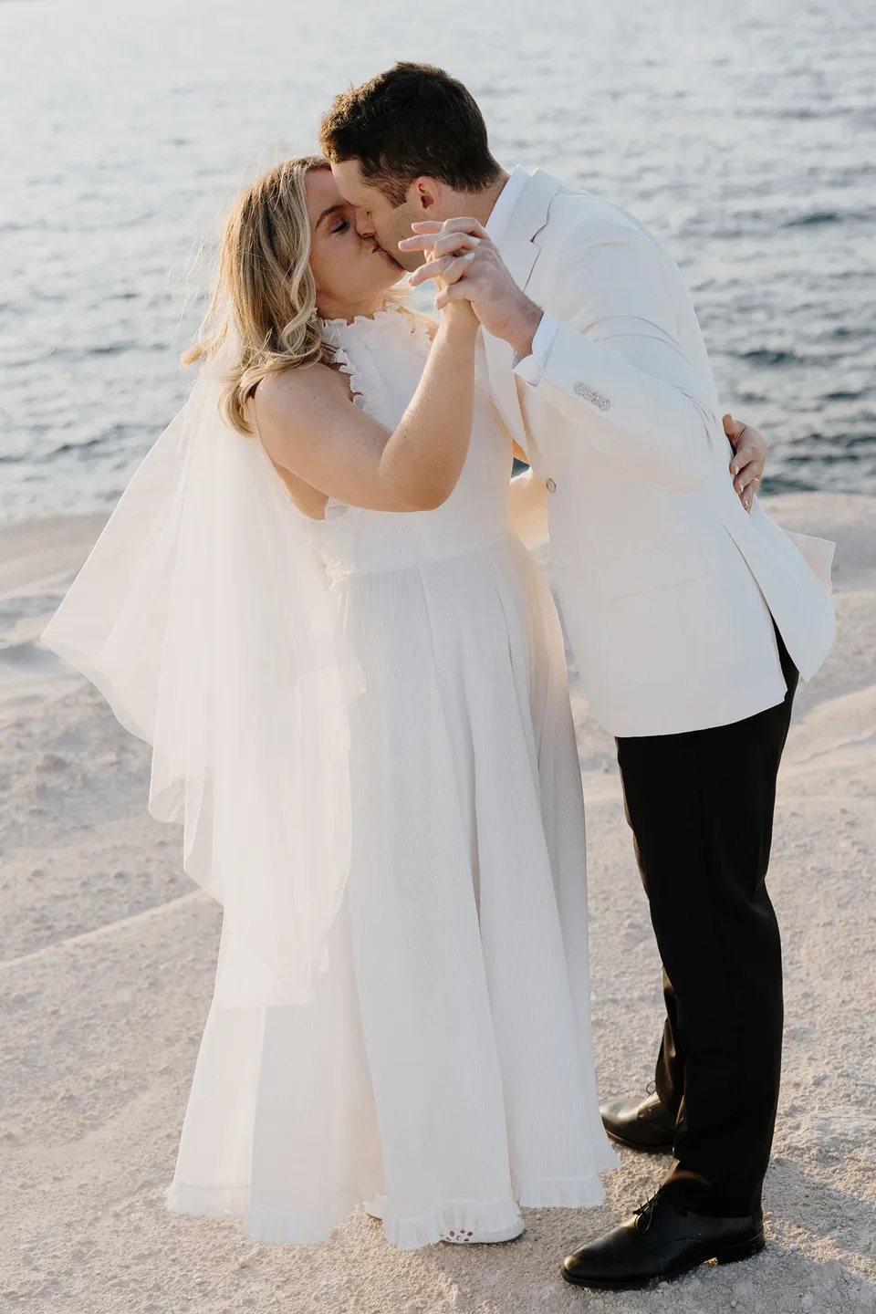 Georgina and Brad kissing after their ceremony, with the blue sea behind them, at Sarakiniko on Milos in the Cyclades, Greece, during an Elopement Collective elopement. Photograph by George Bowden.