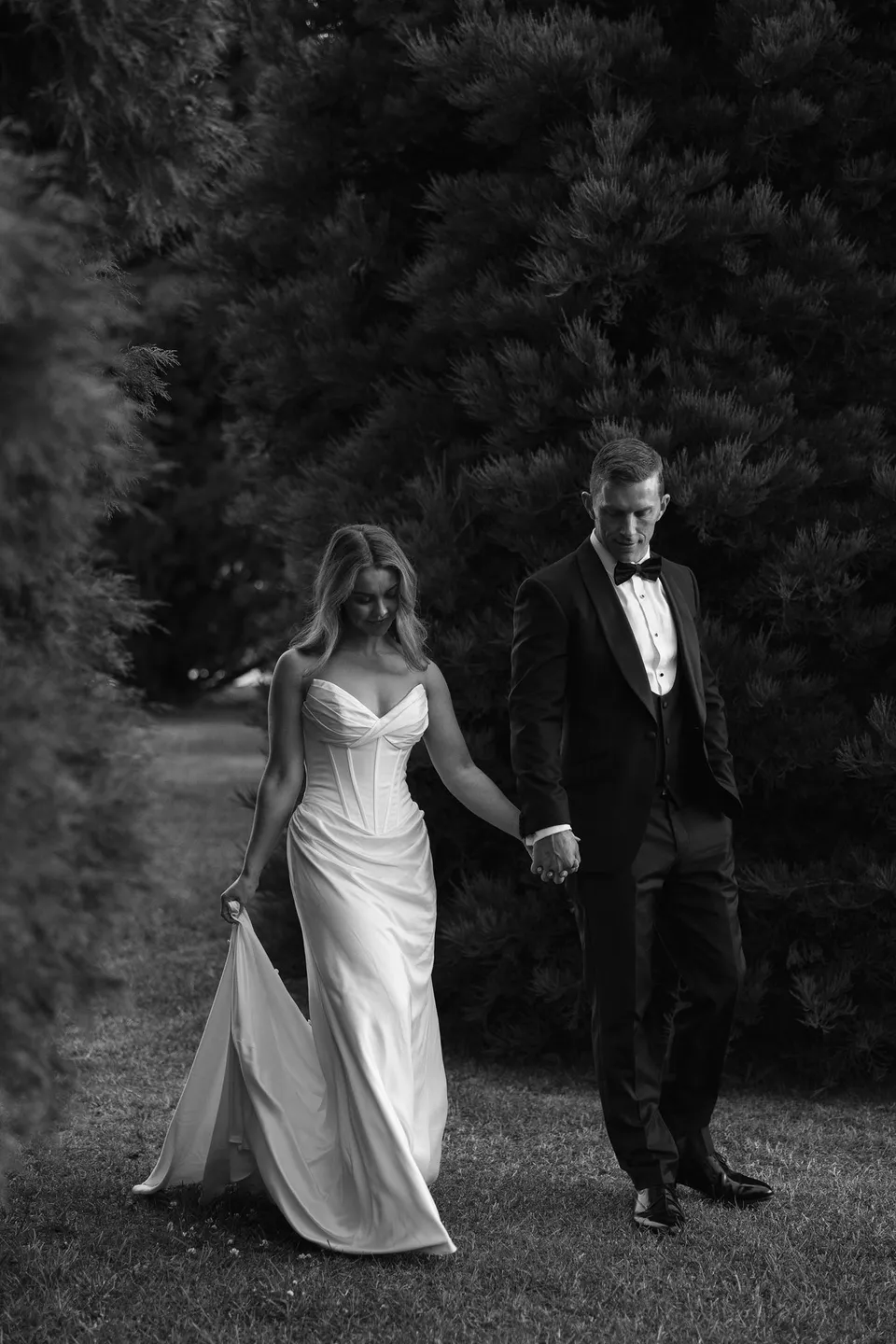 Black and white photograph of Tanesha and Andrew walking hand in hand beneath tall trees, with her dress and veil trailing behind, at Hobart, Tasmania, Australia, for an Elopement Collective summer elopement. Photograph by George Bowden.