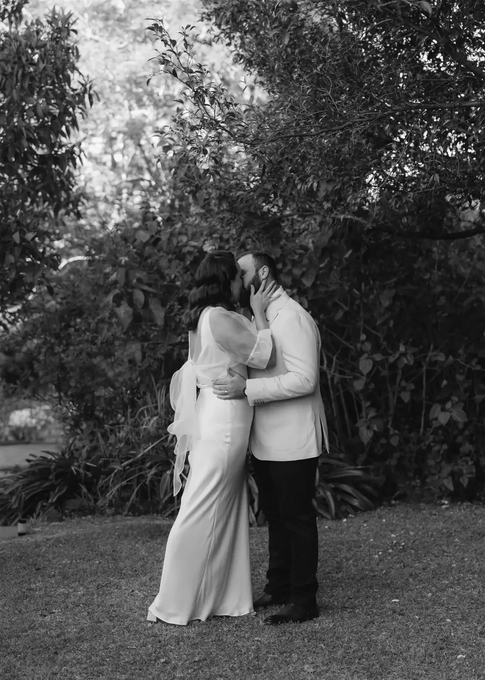 Black-and-white photograph of Tineeka and Chris kissing on the garden lawn, in Montville, Sunshine Coast Hinterland, Queensland, for an Elopement Collective elopement. Photo by Bulb Creative.