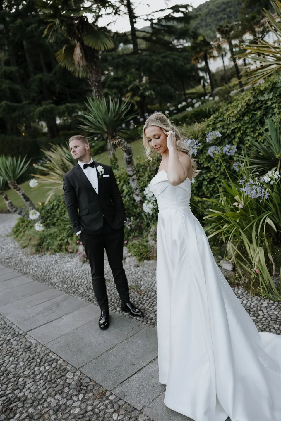 Zoe and Charlton walking together along a garden path, with Zoe lifting a hand to her hair and her white train trailing behind, at Passalacqua in Moltrasio, Lake Como, Italy, for an Elopement Collective elopement. Photo by George Bowden.