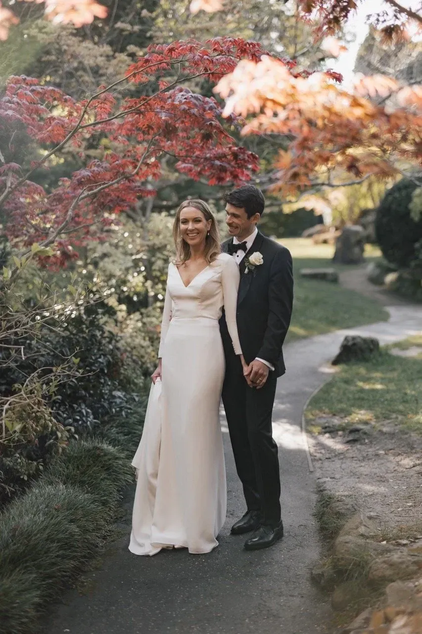A couple hold hands on a narrow garden path beneath colourful foliage.