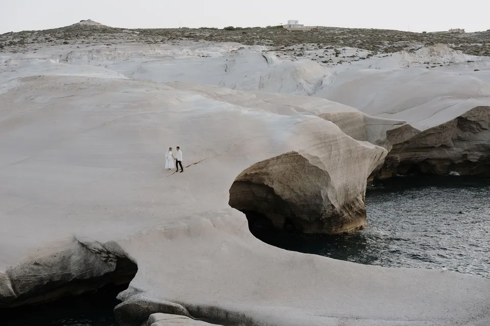 Georgina and Brad holding hands on sculpted white coastal rocks above a broad arched inlet and dark blue sea, at Sarakiniko on Milos in the Cyclades, Greece, during an Elopement Collective elopement. Photograph by George Bowden.