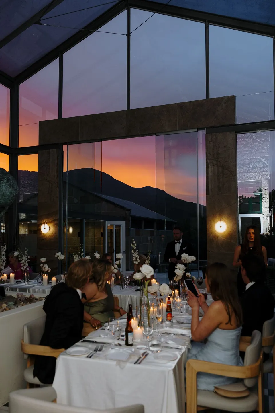 Friends and family seated at a reception table styled by Artaud & Co, with white flowers by The Romantics and sunset colours beyond the glass conservatory, at Islington Hotel in South Hobart, Tasmania, Australia, for an Elopement Collective summer elopement. Photograph by George Bowden.