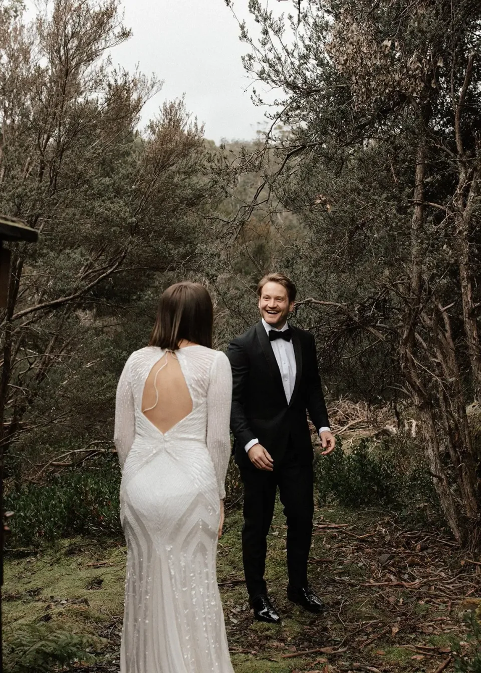 April approaching Josh along a mossy woodland path as he smiles at her, at Cradle Mountain Lodge in Cradle Mountain, Tasmania, for an Elopement Collective elopement. Photo by Bulb Creative.