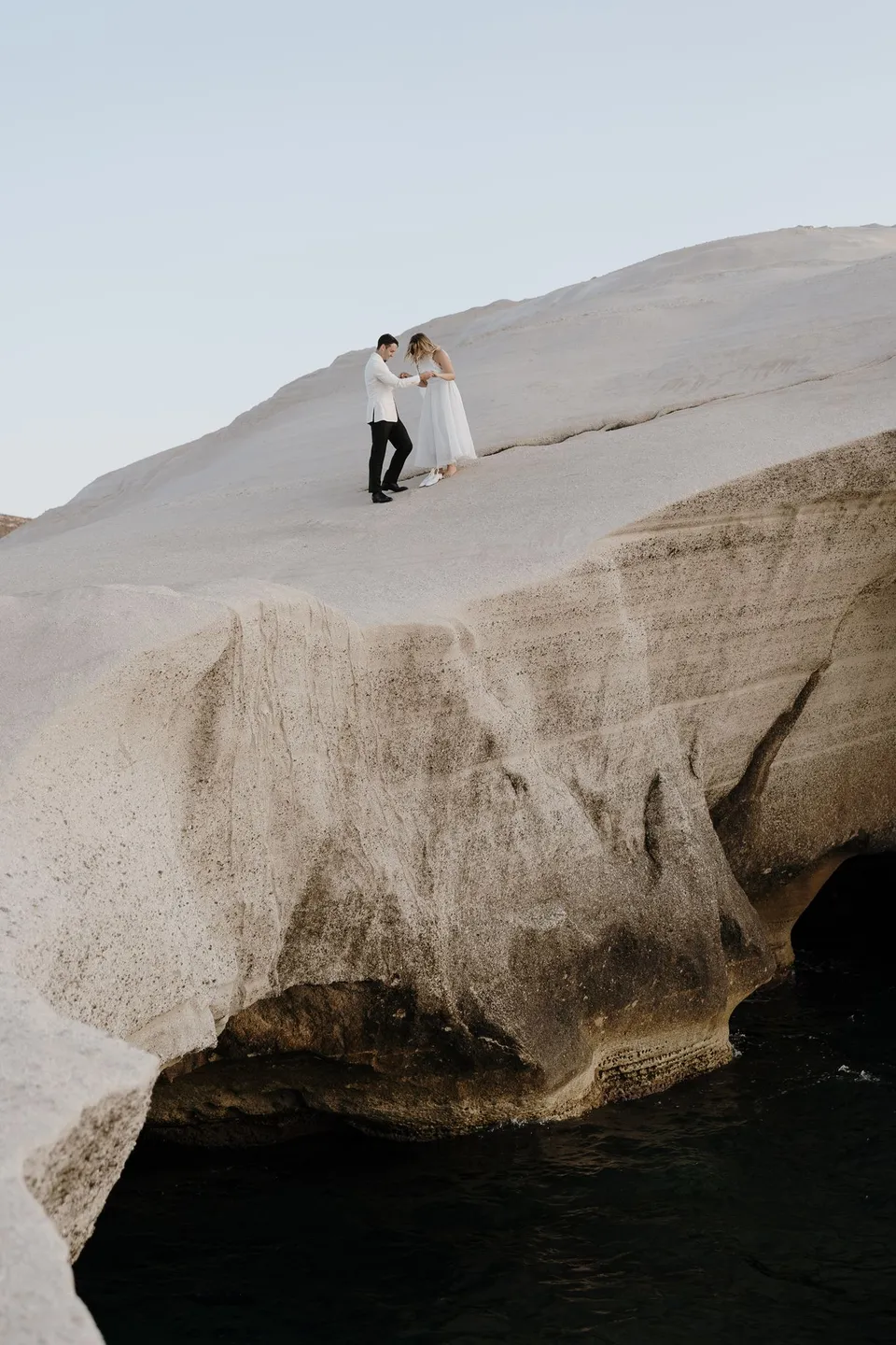 Georgina and Brad standing together on a white cliff slope above blue water and dark shoreline rocks, at Sarakiniko on Milos in the Cyclades, Greece, during an Elopement Collective elopement. Photograph by George Bowden.