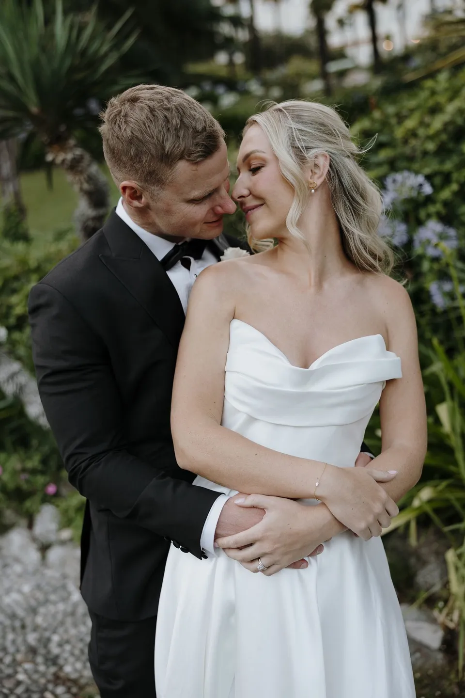 Charlton embracing Zoe from behind as they turn towards each other, with Zoe’s softly waved hair and makeup by Valentina of Sunshine Brides, at Passalacqua in Moltrasio, Lake Como, Italy, for an Elopement Collective elopement. Photo by George Bowden.