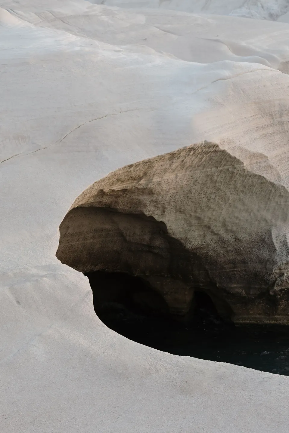 A white cliff headland and rock arch above dark blue water, seen across open pale rocks, at Sarakiniko on Milos in the Cyclades, Greece, during an Elopement Collective elopement. Photograph by George Bowden.