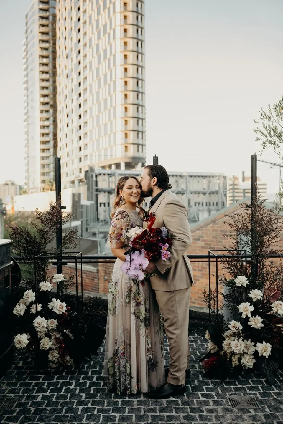 Victoria smiling in Daniel’s embrace among white rooftop flowers by Mrs Gibbons Flowers, at Agnes Restaurant in Fortitude Valley, Brisbane, for an Elopement Collective elopement. Photo by Tessa Shannon.