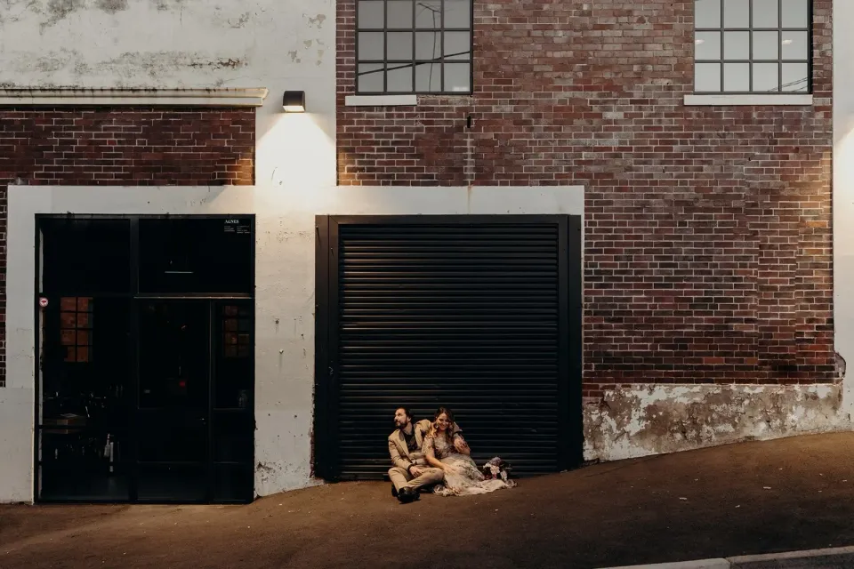 Wide evening portrait of Victoria and Daniel sitting beside a black roller door beneath a brick facade, in Fortitude Valley, Brisbane, for an Elopement Collective elopement. Photo by Tessa Shannon.