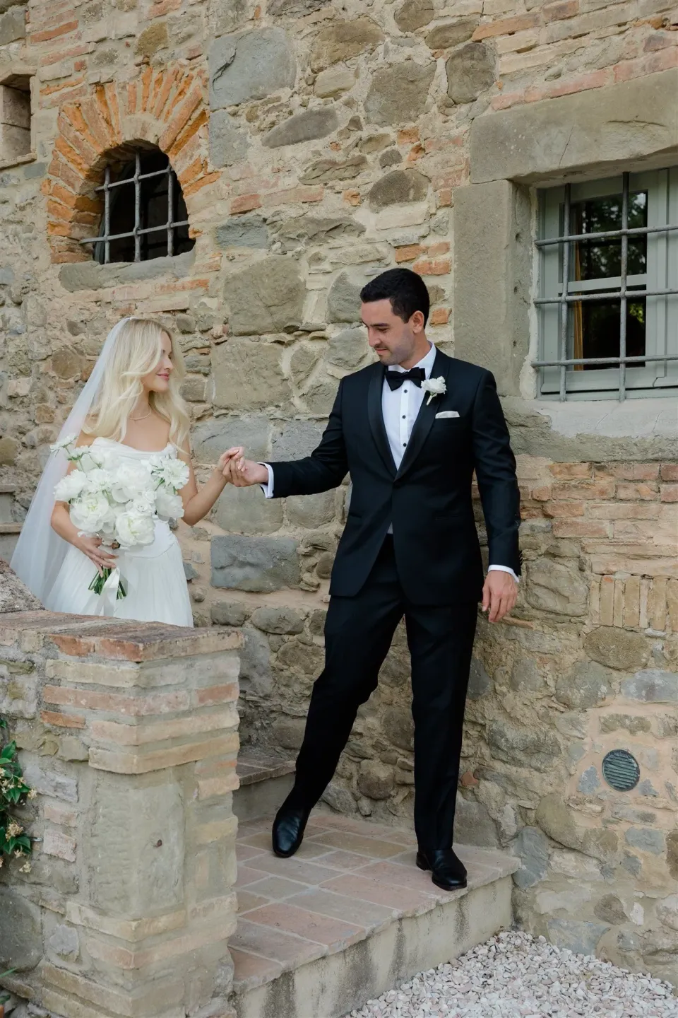 Jack reaching for Shelby’s hand as they step through a stone doorway, with her white bouquet by Le Francesche, in Umbria, Italy, for an Elopement Collective elopement. Photo by James from House of Love.