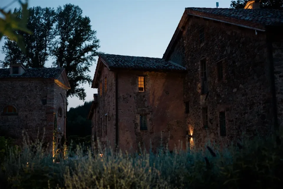 A stone country building glowing with warm window light at dusk beside a quiet garden, in Umbria, Italy, for an Elopement Collective elopement. Photo by James from House of Love.