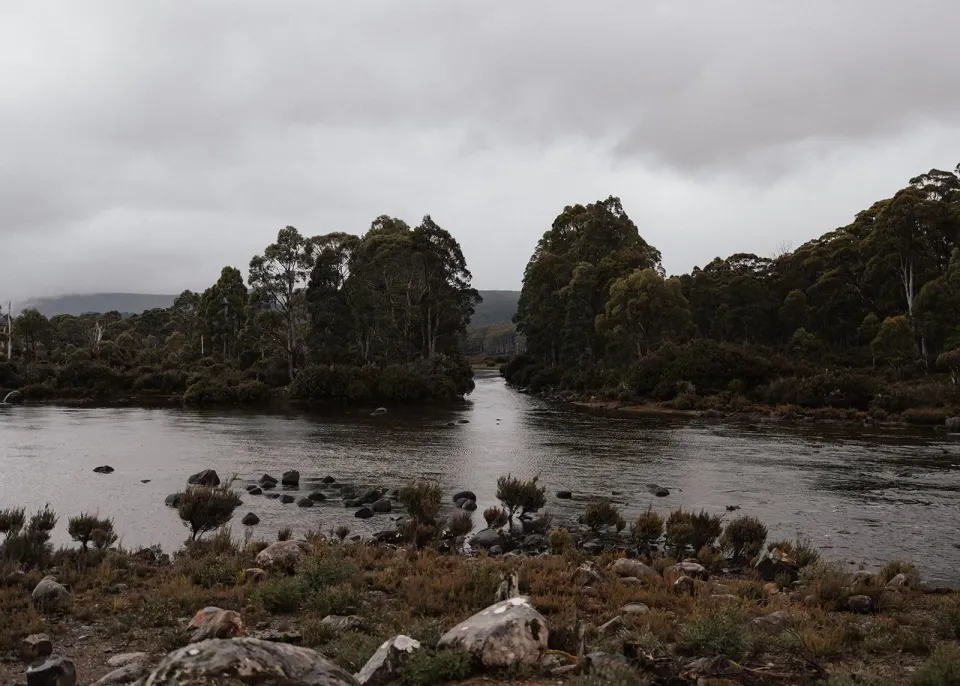 Wide view of Sarah and Jono kissing beside the lake, surrounded by rocks, bush and still water, at Pumphouse Point, Lake St Clair, Tasmania, for an Elopement Collective elopement. Photo by Bulb Creative.