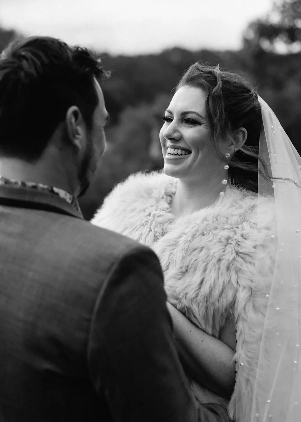 Black-and-white close-up of Sarah smiling up at Jono, her veil falling behind her pale textured wrap, at Pumphouse Point, Lake St Clair, Tasmania, for an Elopement Collective elopement. Photo by Bulb Creative.