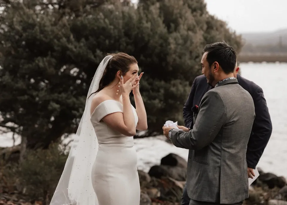 Sarah wiping tears from her face as Jono speaks during their ceremony, with Josh Withers behind them, at Pumphouse Point, Lake St Clair, Tasmania, for an Elopement Collective elopement. Photo by Bulb Creative.