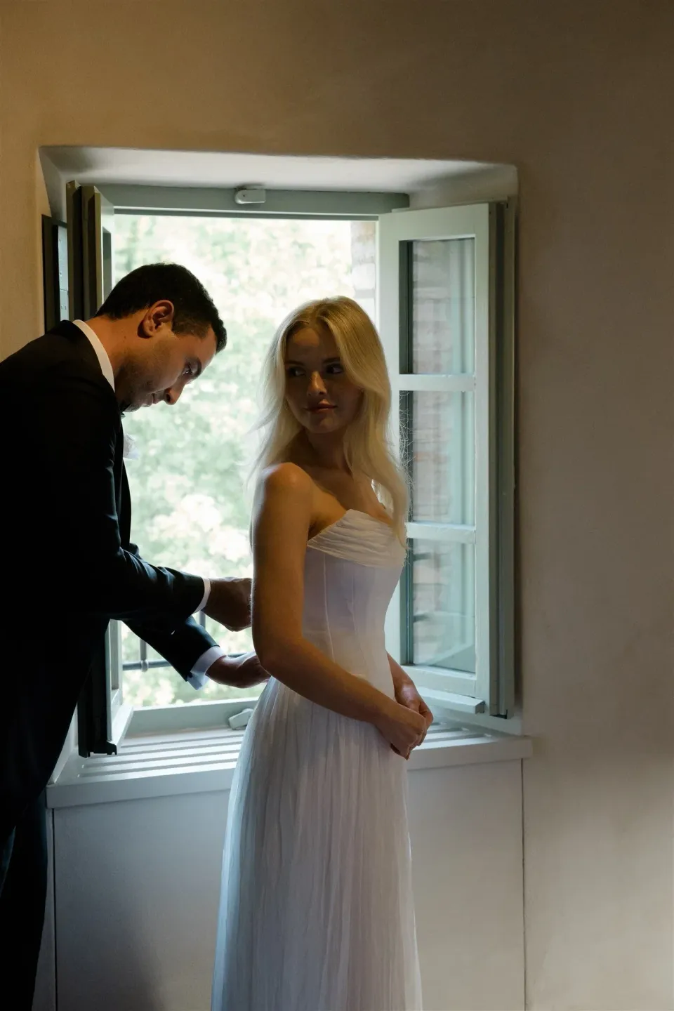 Jack helping Shelby with her wedding dress beside an open window, in Umbria, Italy, for an Elopement Collective elopement. Photo by James from House of Love.