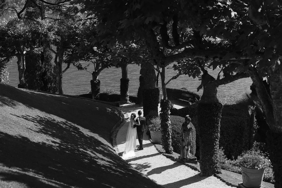 Black and white photograph of Lyndal and Lachlan walking along a garden path beneath sculpted trees at Villa del Balbianello, with shadows across the lawn and lake views beyond, at Lake Como in Lombardy, Italy, for an Elopement Collective elopement. Photograph by George Bowden.