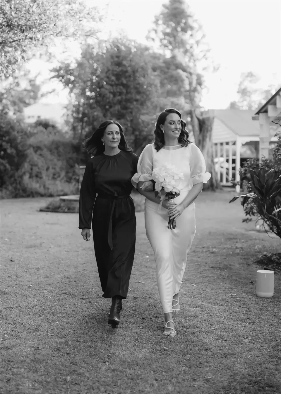 Black-and-white photograph of Tineeka walking beside a woman in a black dress, carrying her bouquet by Fifth Ave, in Montville, Sunshine Coast Hinterland, Queensland, for an Elopement Collective elopement. Photo by Bulb Creative.