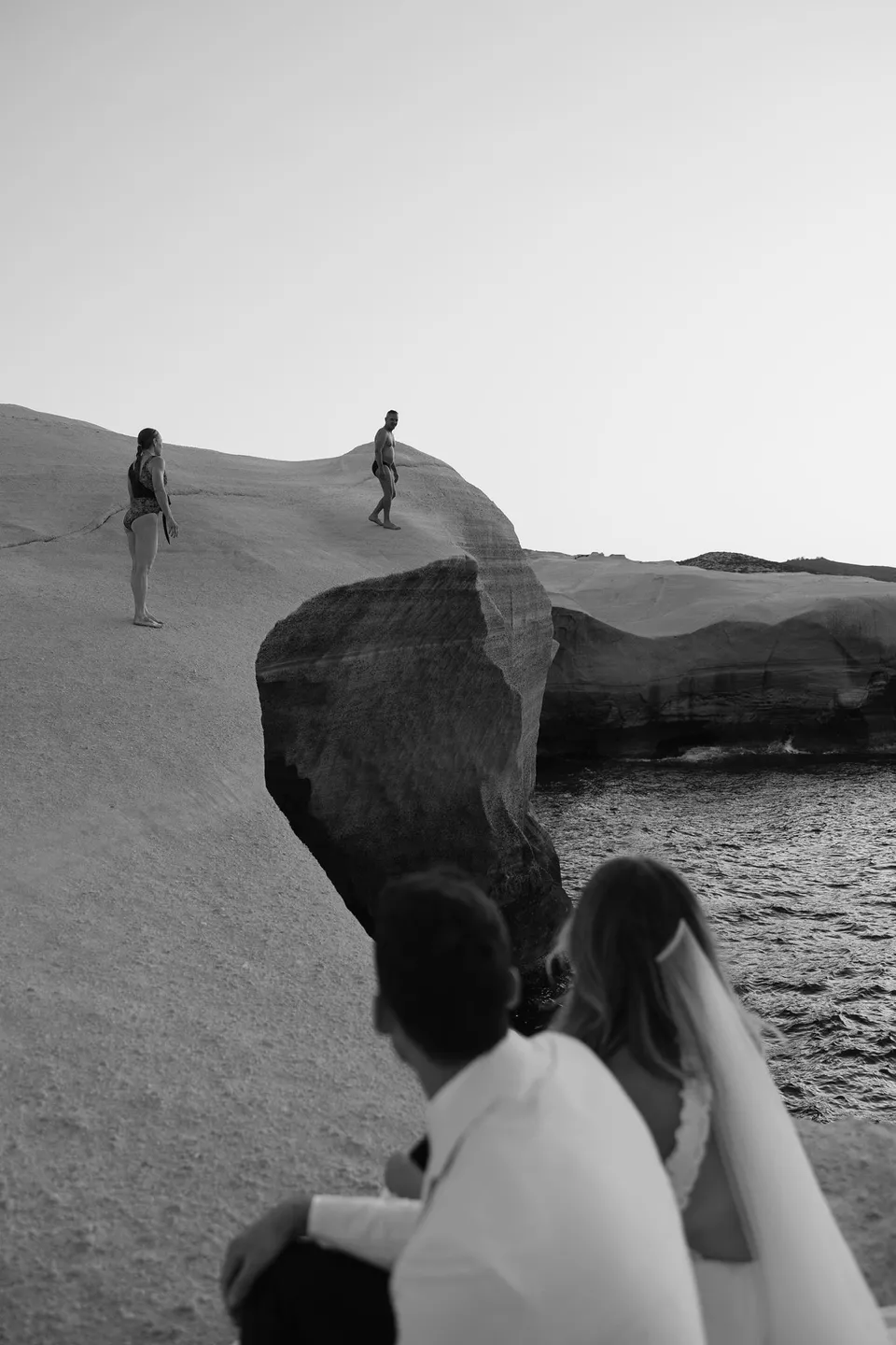 Black and white view of Georgina and Brad looking across an inlet towards two visitors standing on the white rocks, at Sarakiniko on Milos in the Cyclades, Greece, during an Elopement Collective elopement. Photograph by George Bowden.