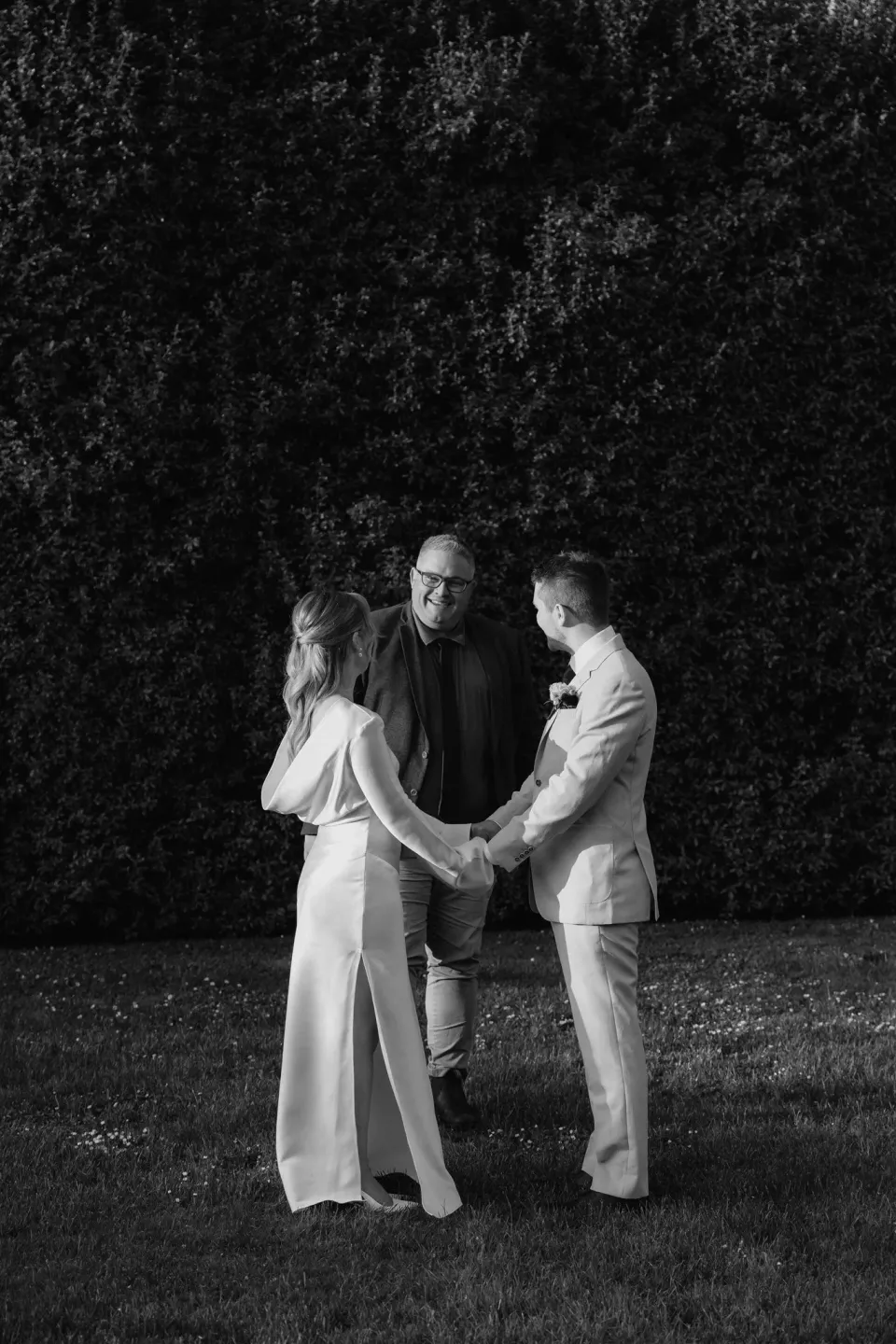 Black-and-white photograph of Maddy and Cameron holding hands during their garden ceremony with Josh Withers, at The Mayor’s Residence in Daylesford, Victoria, for an Elopement Collective elopement. Photo by Bulb Creative.