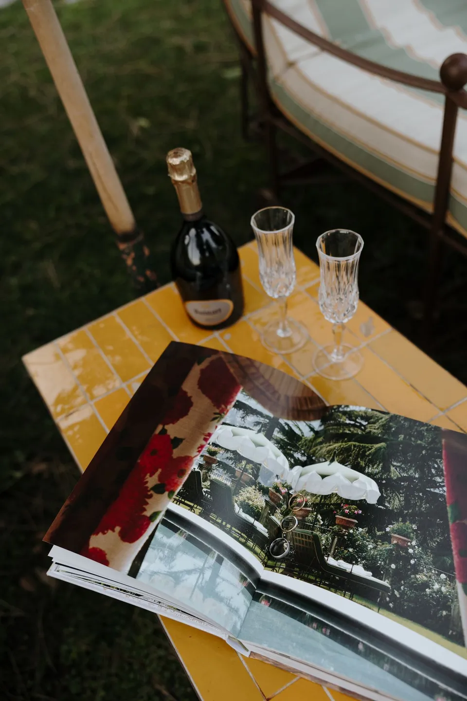 A champagne bottle and two filled flutes beside an open book about Passalacqua on a yellow table among garden chairs, at Passalacqua in Moltrasio, Lake Como, Italy, for an Elopement Collective elopement. Photo by George Bowden.