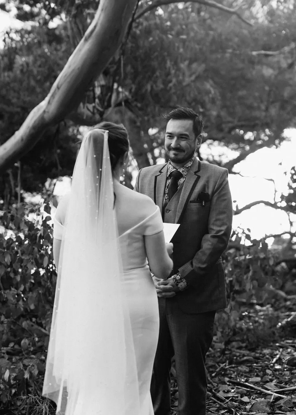 Black-and-white photograph of Jono smiling at Sarah during their ceremony beneath the trees, at Pumphouse Point, Lake St Clair, Tasmania, for an Elopement Collective elopement. Photo by Bulb Creative.