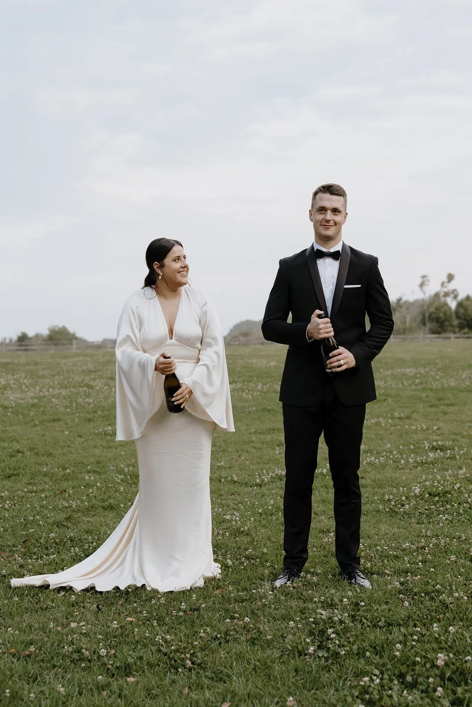 Laura and Matthew preparing to open a bottle of sparkling wine in a paddock. At The Farm and Three Blue Ducks in Ewingsdale, Byron Bay, for an Elopement Collective elopement. Photograph by George Bowden.