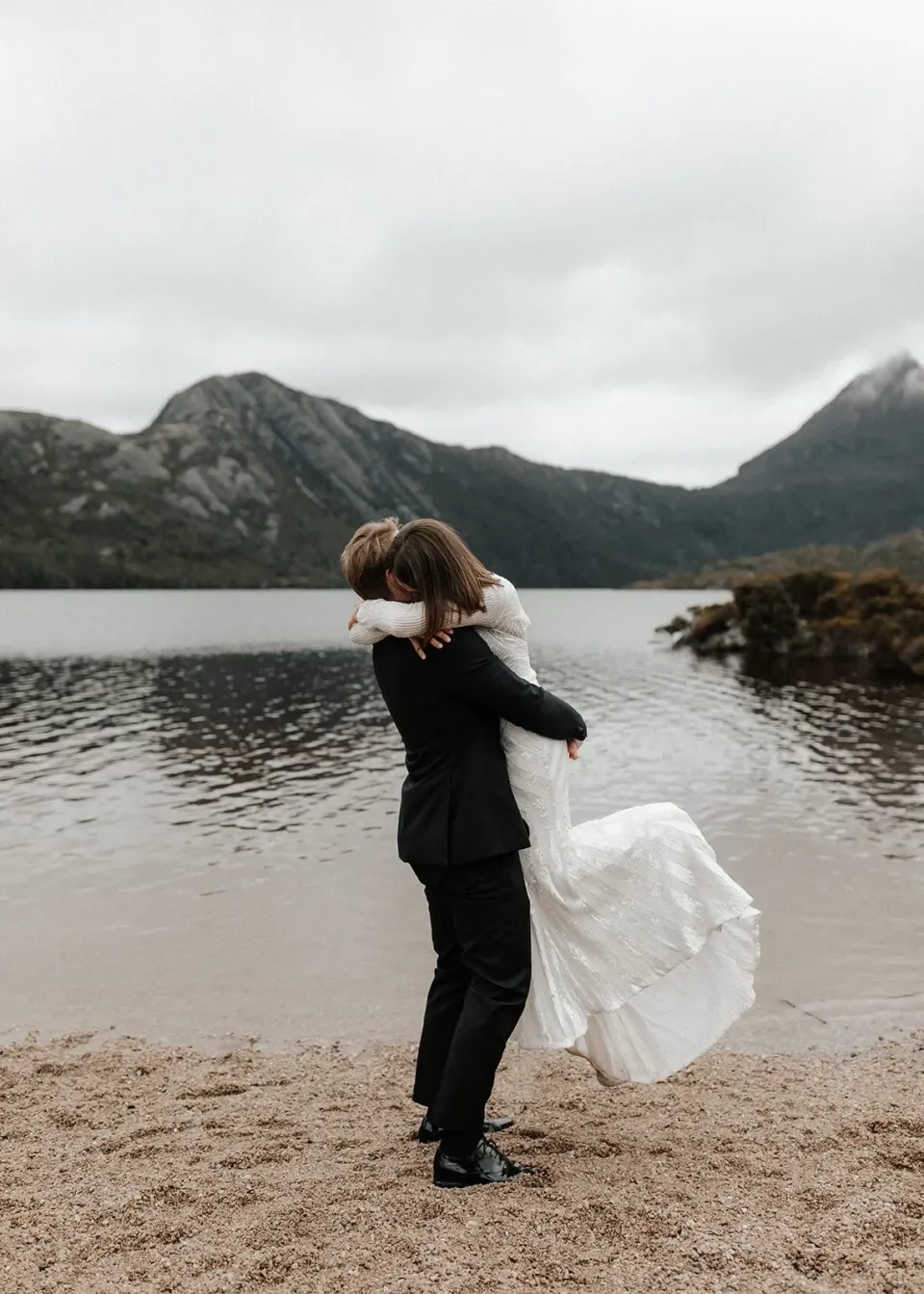 Josh lifting April into an embrace, her dress sweeping above the lakeshore, at Dove Lake, Cradle Mountain, Tasmania, for an Elopement Collective elopement. Photo by Bulb Creative.