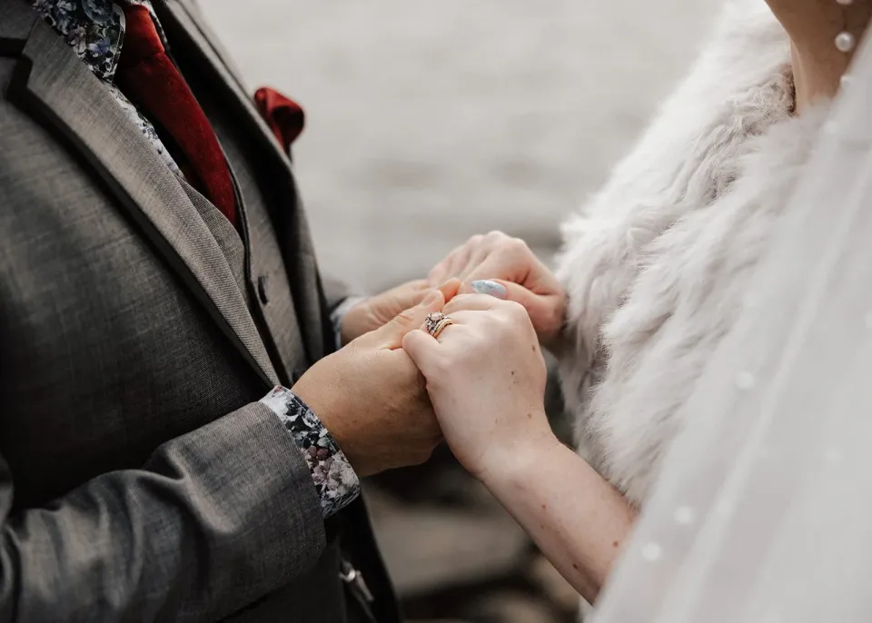 Close-up of Sarah and Jono holding hands, with their wedding rings, his grey suit and her textured wrap visible, at Pumphouse Point, Lake St Clair, Tasmania, for an Elopement Collective elopement. Photo by Bulb Creative.