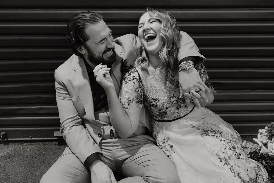 Black-and-white portrait of Victoria laughing with Daniel’s arm around her shoulders while seated outdoors, in Fortitude Valley, Brisbane, for an Elopement Collective elopement. Photo by Tessa Shannon.