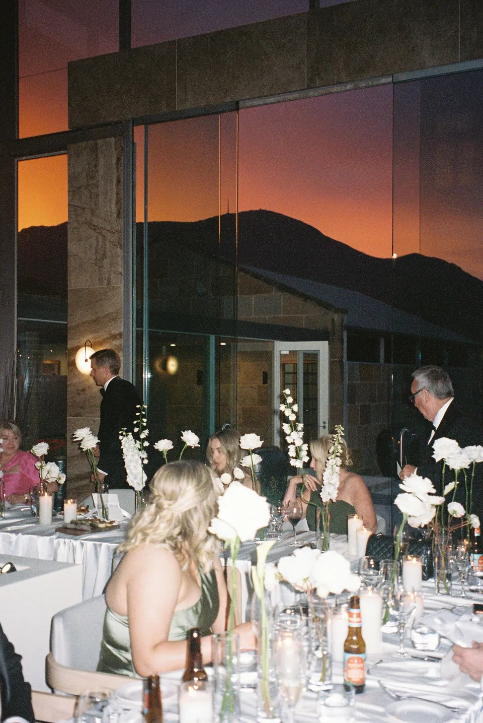 Guests gathered around tables styled by Artaud & Co with white flowers by The Romantics, while the orange sunset frames the mountain through the conservatory windows, at Islington Hotel in South Hobart, Tasmania, Australia, for an Elopement Collective summer elopement. Photograph by George Bowden.