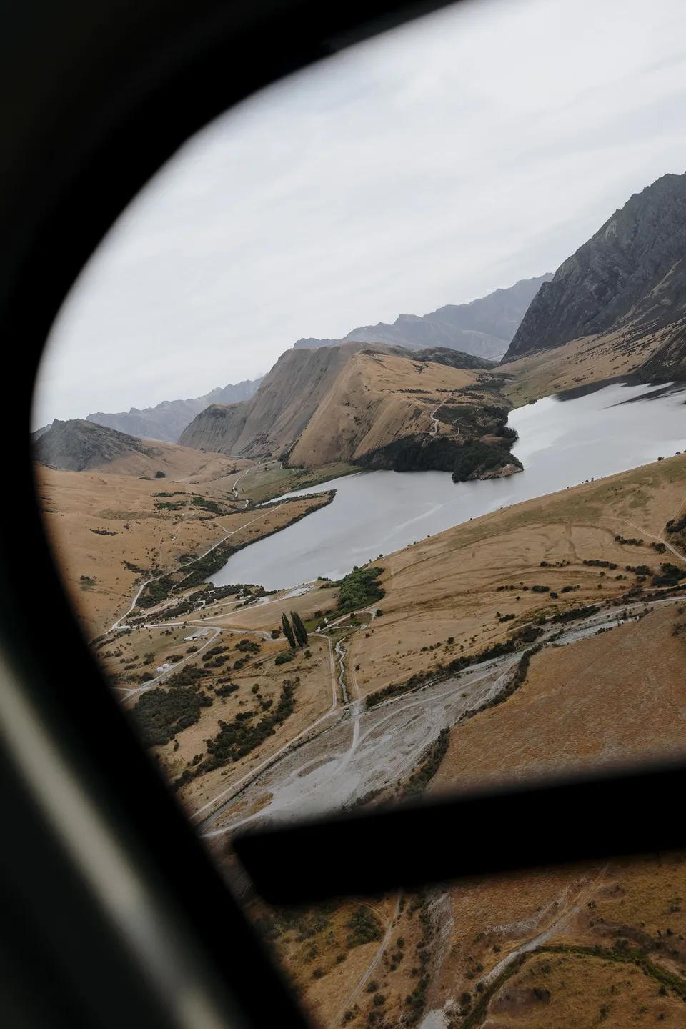 A mountain lake, winding track and golden slopes seen through the helicopter window during the flight to their alpine portraits, above Queenstown, New Zealand, for an Elopement Collective Queenstown elopement. Photo by George Bowden.