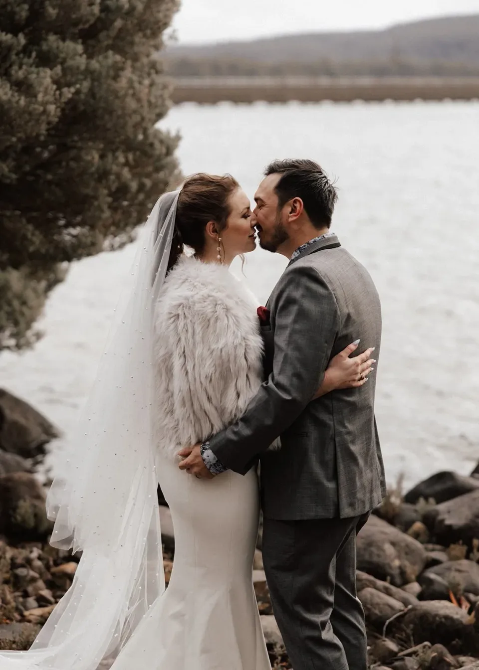 Sarah and Jono looking at each other beside the lake, her pale textured wrap around her shoulders, at Pumphouse Point, Lake St Clair, Tasmania, for an Elopement Collective elopement. Photo by Bulb Creative.