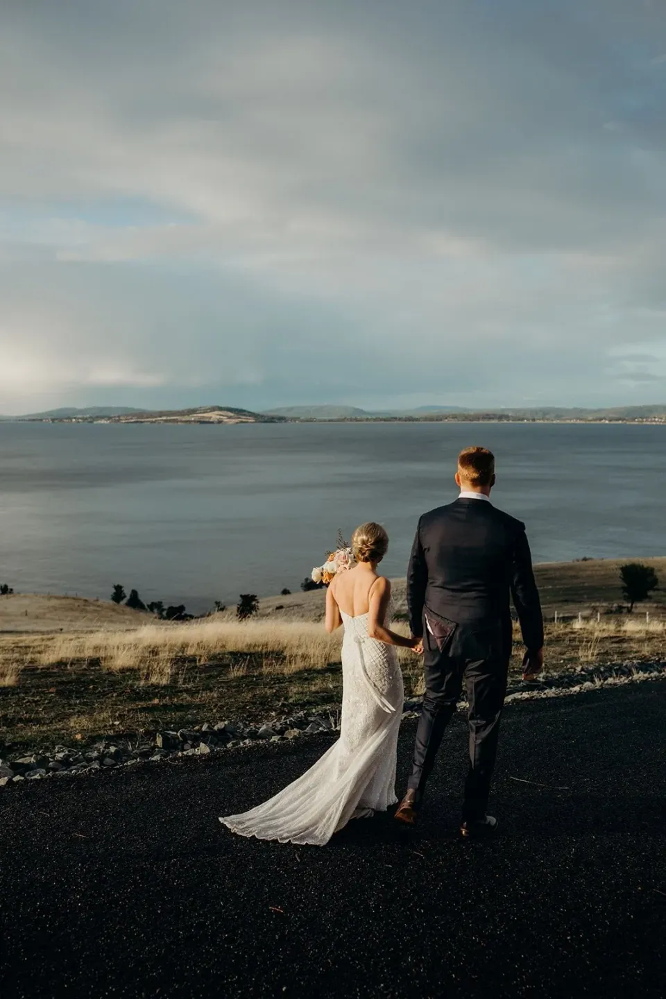 A couple look across a bay from a hillside, the bride's train spread behind her.