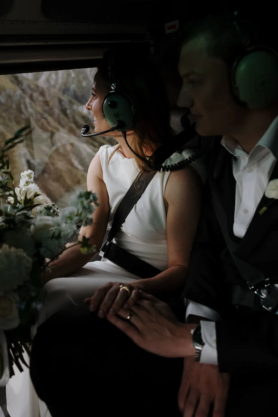 Neve and Hamish wearing headsets and holding hands inside the helicopter, with Neve’s Orange Blossom bouquet on her lap, above Queenstown, New Zealand, for an Elopement Collective Queenstown elopement. Photo by George Bowden.