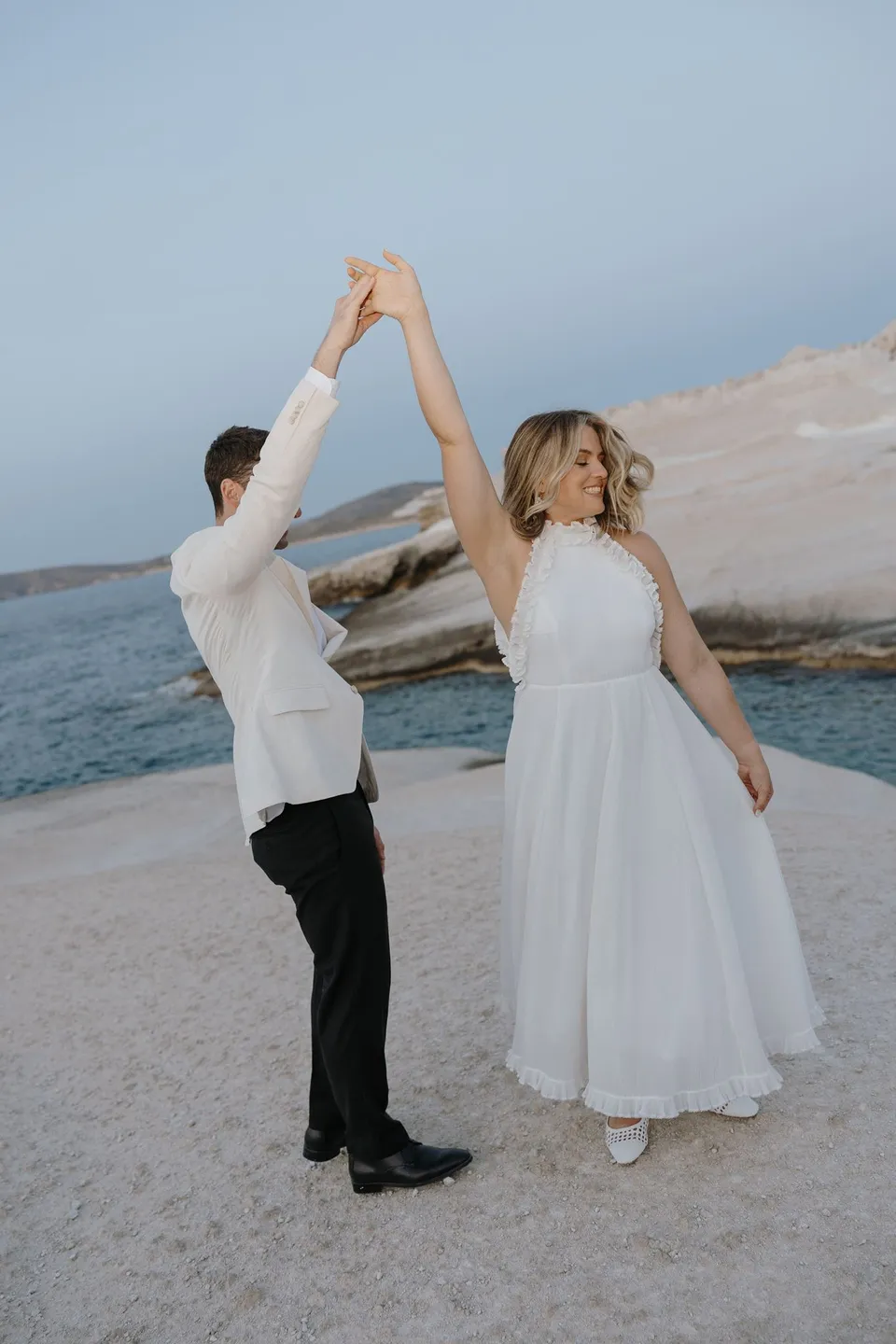Brad raising Georgina’s hand to twirl her beside the sea in the evening light, at Sarakiniko on Milos in the Cyclades, Greece, during an Elopement Collective elopement. Photograph by George Bowden.