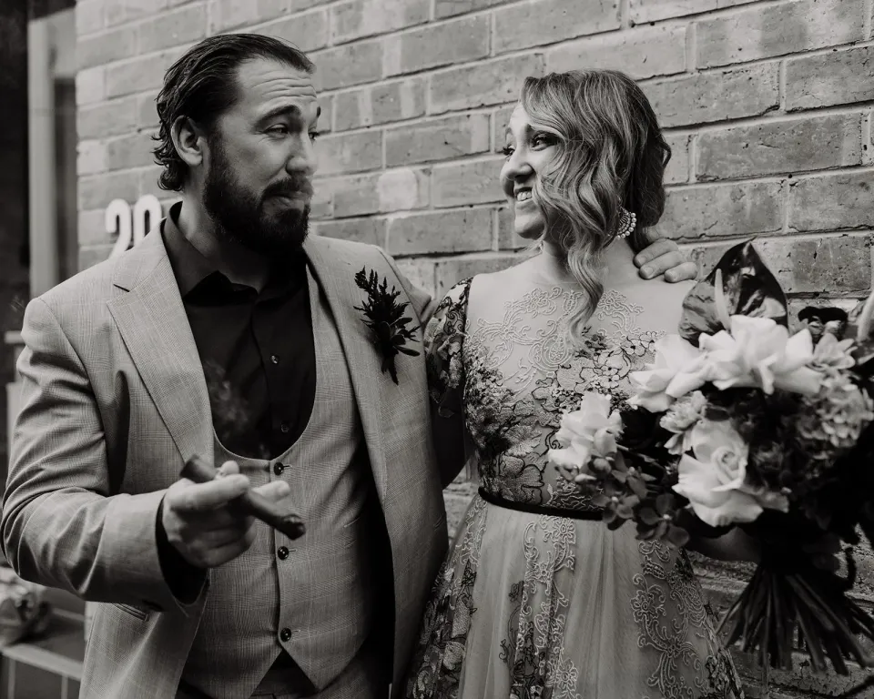 Black-and-white portrait of Daniel with a cigar beside Victoria and her bouquet by Mrs Gibbons Flowers, in Fortitude Valley, Brisbane, for an Elopement Collective elopement. Photo by Tessa Shannon.