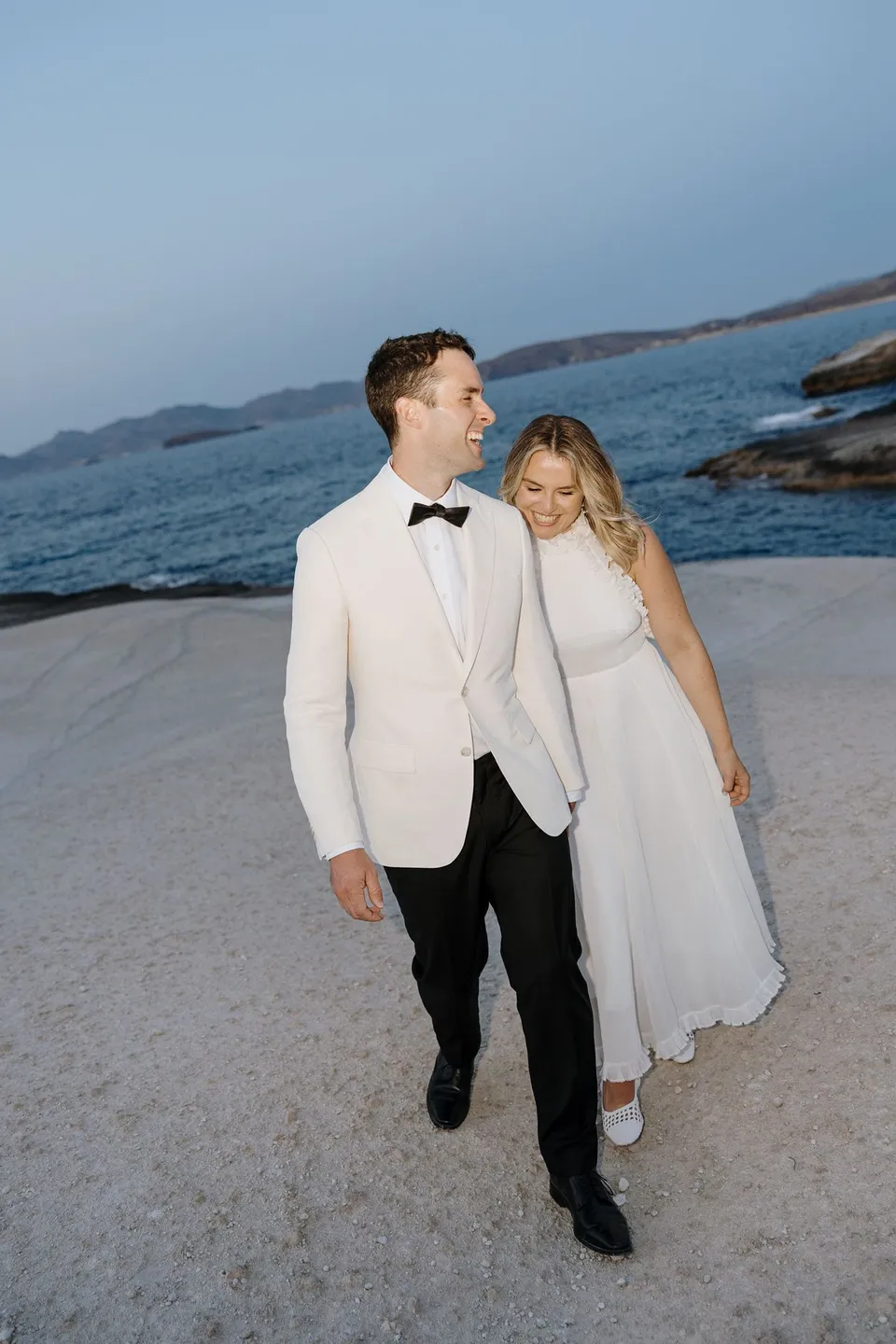 Georgina resting her head on Brad’s shoulder as they walk together across the rocks, at Sarakiniko on Milos in the Cyclades, Greece, during an Elopement Collective elopement. Photograph by George Bowden.