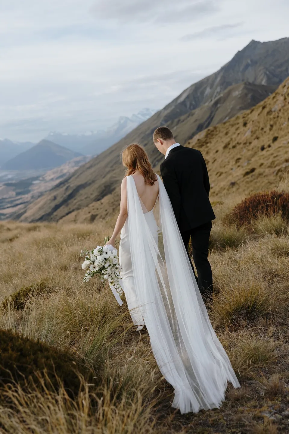 Neve and Hamish walking through alpine tussock, with Neve carrying her Orange Blossom bouquet and her sheer cape flowing behind her, on Big Geordie above Queenstown, New Zealand, for an Elopement Collective Queenstown elopement. Photo by George Bowden.