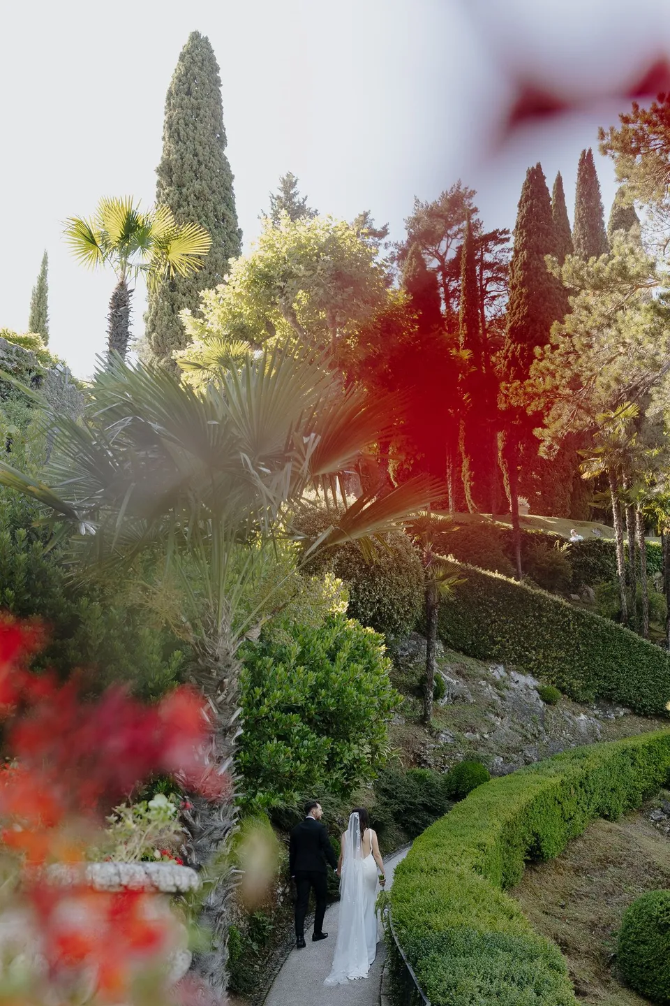 Lyndal and Lachlan walking along a path between clipped hedges, palms and cypress trees at Villa del Balbianello, seen through soft red flowers in the foreground, at Lake Como in Lombardy, Italy, for an Elopement Collective elopement. Photograph by George Bowden.