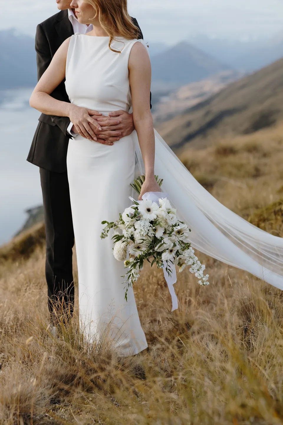 Hamish holding Neve around the waist as she carries her white Orange Blossom bouquet, with her sheer cape moving across the mountain grass, on Big Geordie above Queenstown, New Zealand, for an Elopement Collective Queenstown elopement. Photo by George Bowden.
