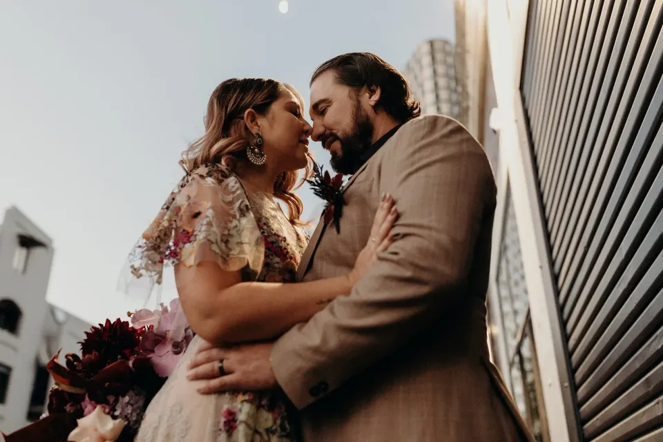 Low-angle portrait of Victoria and Daniel leaning close beside a black roller door, with her bouquet by Mrs Gibbons Flowers, in Fortitude Valley, Brisbane, for an Elopement Collective elopement. Photo by Tessa Shannon.