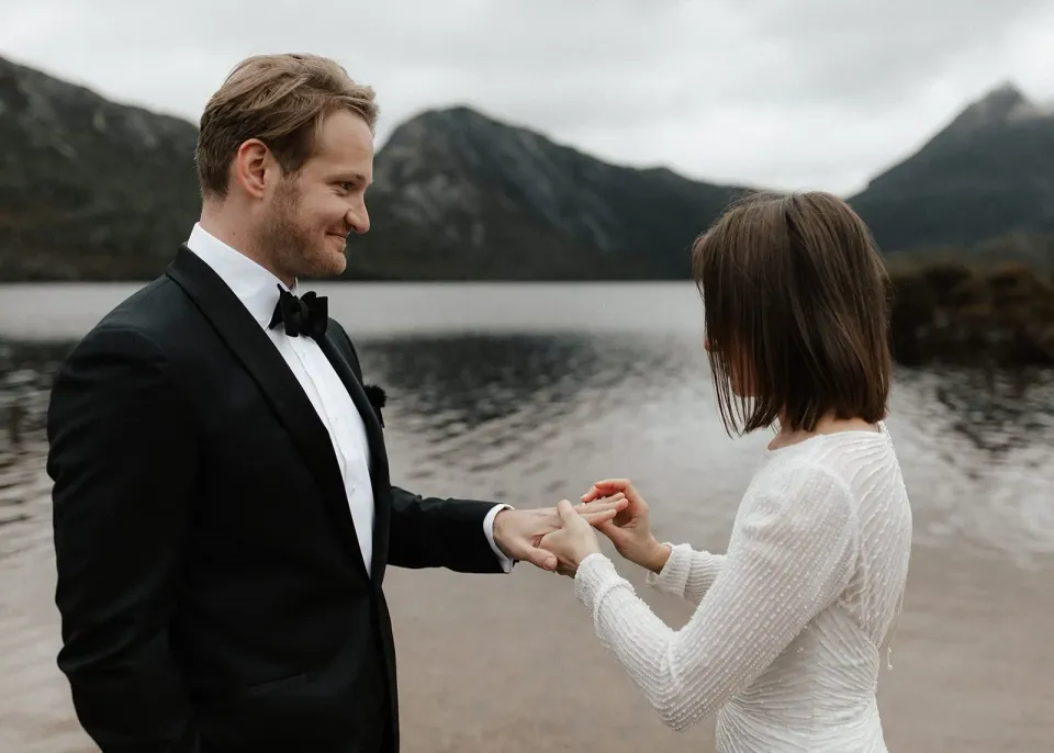 April placing a wedding ring on Josh’s hand beside the lake, at Dove Lake, Cradle Mountain, Tasmania, for an Elopement Collective elopement. Photo by Bulb Creative.