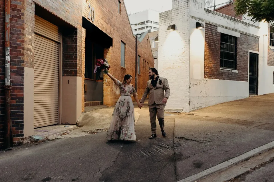 Victoria raising her bouquet by Mrs Gibbons Flowers as she and Daniel walk hand in hand down a city street, in Fortitude Valley, Brisbane, for an Elopement Collective elopement. Photo by Tessa Shannon.