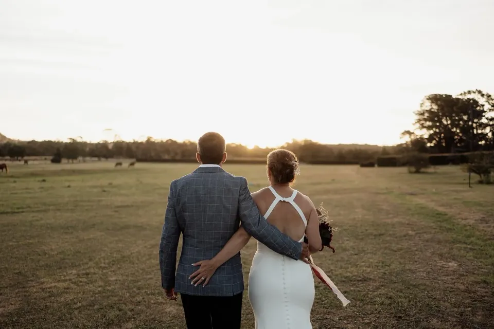 A couple walk arm in arm through an open paddock in the evening light.